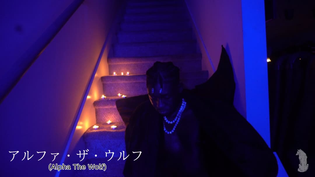 Alpha The Wolf crouched at the foot of a candlelit staircase wrapped in a large dark cape, pearl bead necklace visible at the chest, blue UV light flooding the hall, the Japanese text overlay reading his name adding a cinematic title-card quality