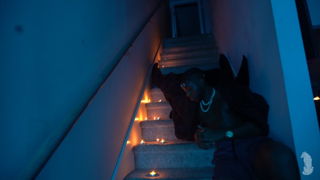 Alpha The Wolf seated low on a candlelit staircase, body angled downward and one hand extended toward the lowest step, pearl necklace and watch catching the warm flame glow against a deep blue ambient fill