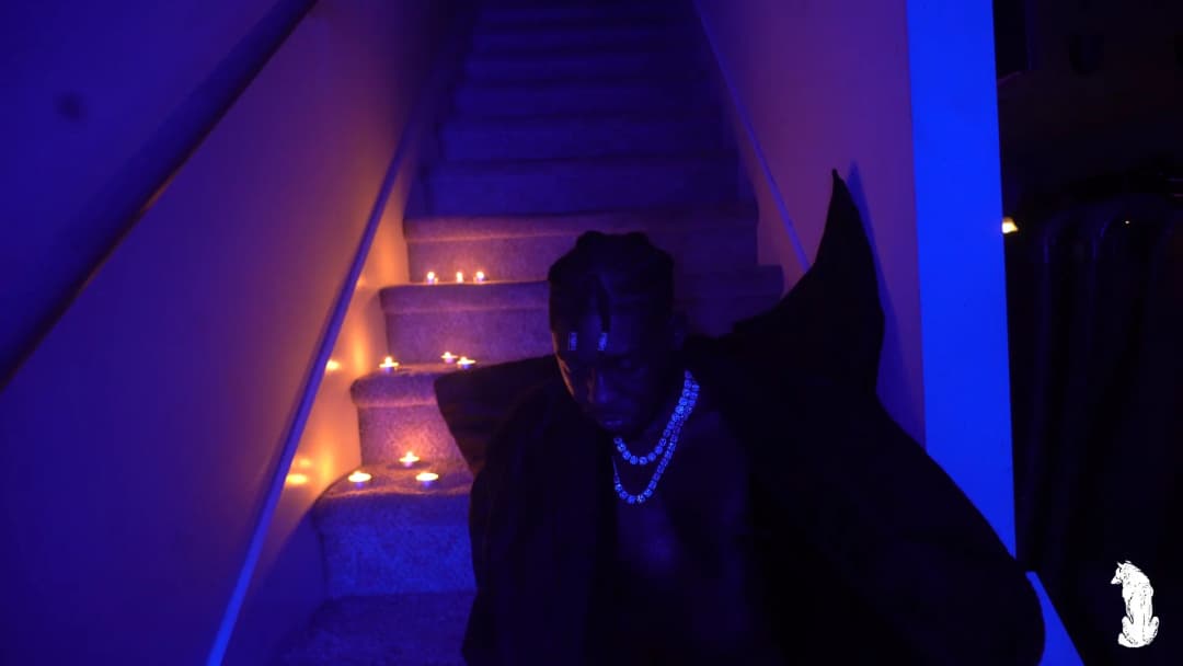 Alpha The Wolf seated at the base of the candlelit stairs in a wide frontal shot, cape spread to either side, iced-out chain glowing brilliantly under UV blue light as candlelight climbs the steps behind him