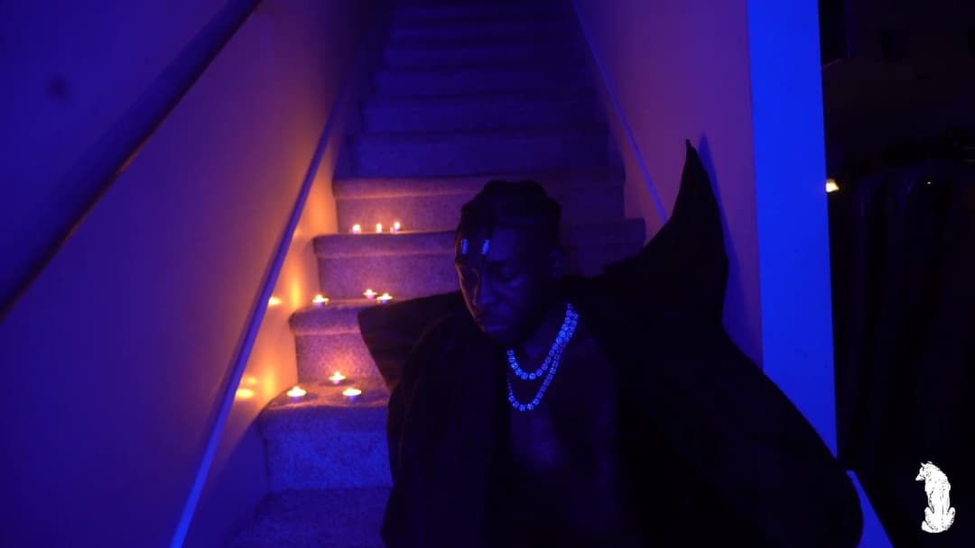 Alpha The Wolf seated at the foot of the candle-lined stairs facing forward, dark cape framing his silhouette, iced chain glowing under blue UV, eyes downcast in quiet introspection as warm candlelight climbs the steps behind him