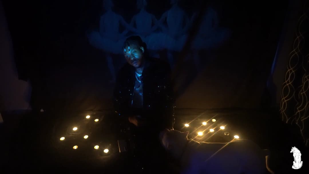 Alpha The Wolf standing between two clusters of candles arranged on the dark floor, facing the camera with chain and pearl necklace glowing under blue light, a projected mural of figures looming on the wall high behind him