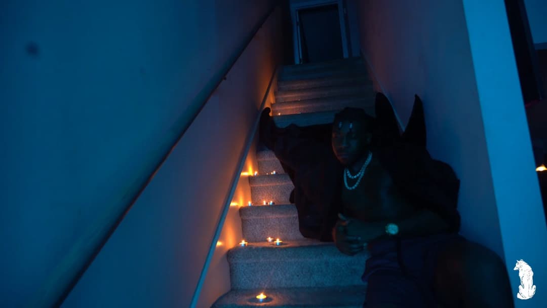 Alpha The Wolf seated on the candlelit stairs leaning forward, pearl necklace hanging, gaze directed toward the camera with relaxed ease, warm candle glow framing his lower body against the cool blue of the stairwell above