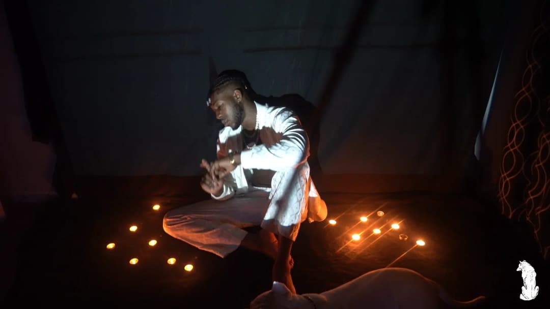 Alpha The Wolf crouched low at the center of the candlelit floor in white, hands moving expressively in front of him, candlelight casting warm highlights across his torso and face in an engaged, conversational posture