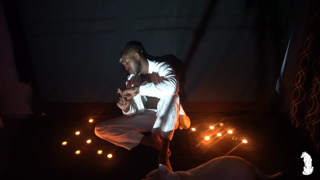 Alpha The Wolf seated among the candles in white, leaning slightly forward with both hands gesturing open-palmed, the candle arrangement fanning out on both sides, the background draped in dark fabric