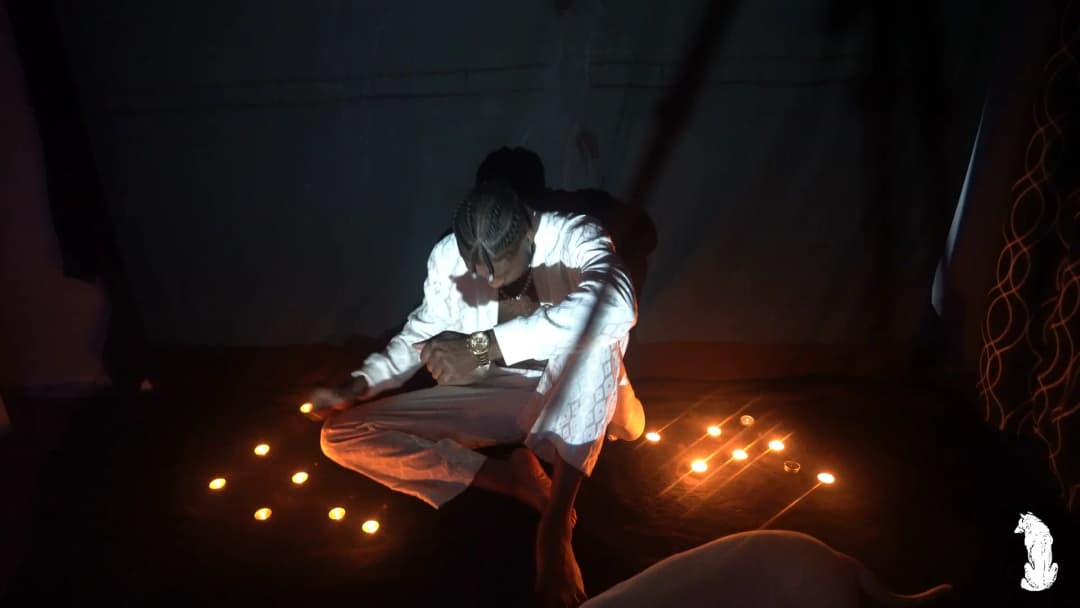 Alpha The Wolf crouched over the candle ring in white, head bowed almost to the floor with both hands flat near the flames, the shot pulled back to reveal the full scale of the candle arrangement spreading across the dark floor around him