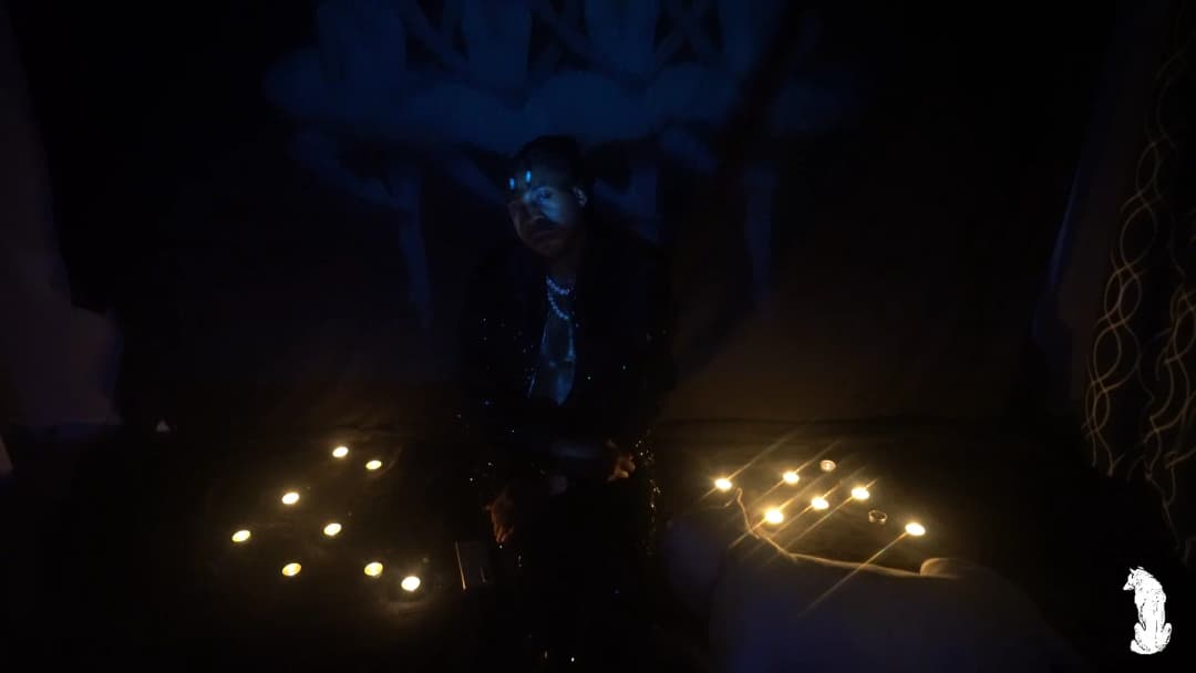 Alpha The Wolf standing between the twin candle clusters, face tilted upward under blue UV light, chain necklace glowing at his chest, the projected mural of figures visible high on the back wall in this wide, ritualistic framing