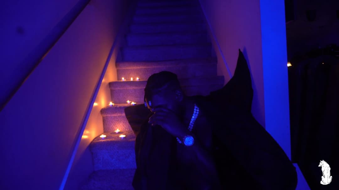 Alpha The Wolf at the bottom of the candle-lit staircase, hand raised to his chin in a contemplative gesture, blue iced watch and chain catching UV light, cape gathered around him as he faces the camera with a guarded expression