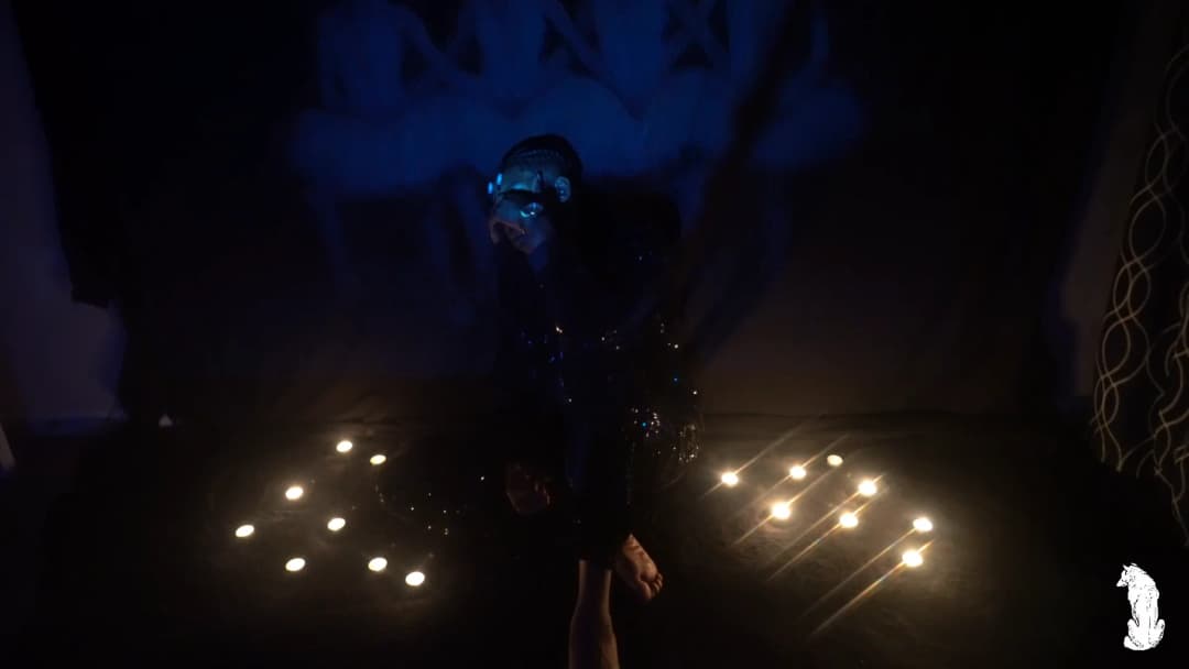 Alpha The Wolf standing in the candle circle in a dark sequined jacket that catches specks of blue light, face slightly obscured as he looks down and to the side, the mural projection looming large on the wall behind him