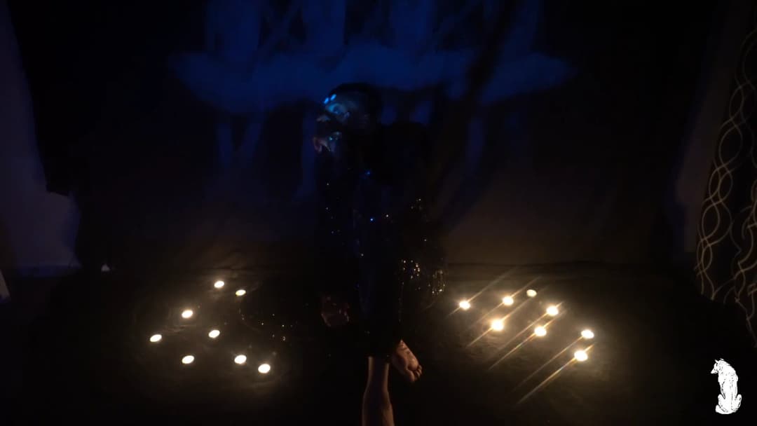 Alpha The Wolf standing in the candle circle, head tilted back slightly under blue UV illumination with one hand raised near his face, the reflective jacket catching scattered points of light, the scene hazy with atmosphere