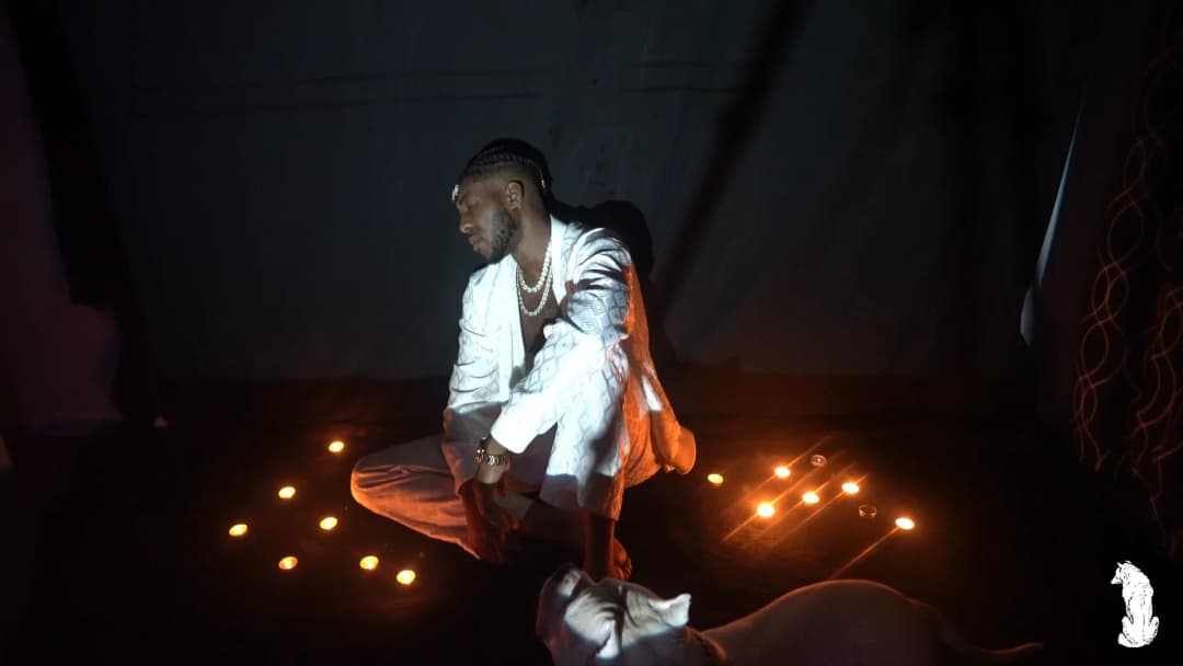 Alpha The Wolf seated in the white outfit among the floor candles, weight shifted to one side and gaze forward, the candlelight soft and warm at his feet while the background fades to near-total black on either side