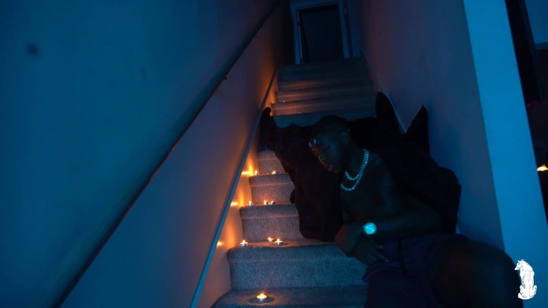 Alpha The Wolf seated low on the candlelit staircase with his back largely to the camera, leaning sideways with one hand gripping the railing, pearl necklace visible from the side under the cool blue ambient light