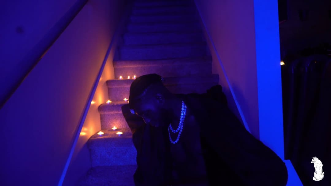 Alpha The Wolf at the foot of the candlelit staircase, facing front with head lowered and hand raised near his cheek, iced chain and watch both catching the blue UV light, cape spread wide as candles flicker on the steps behind him