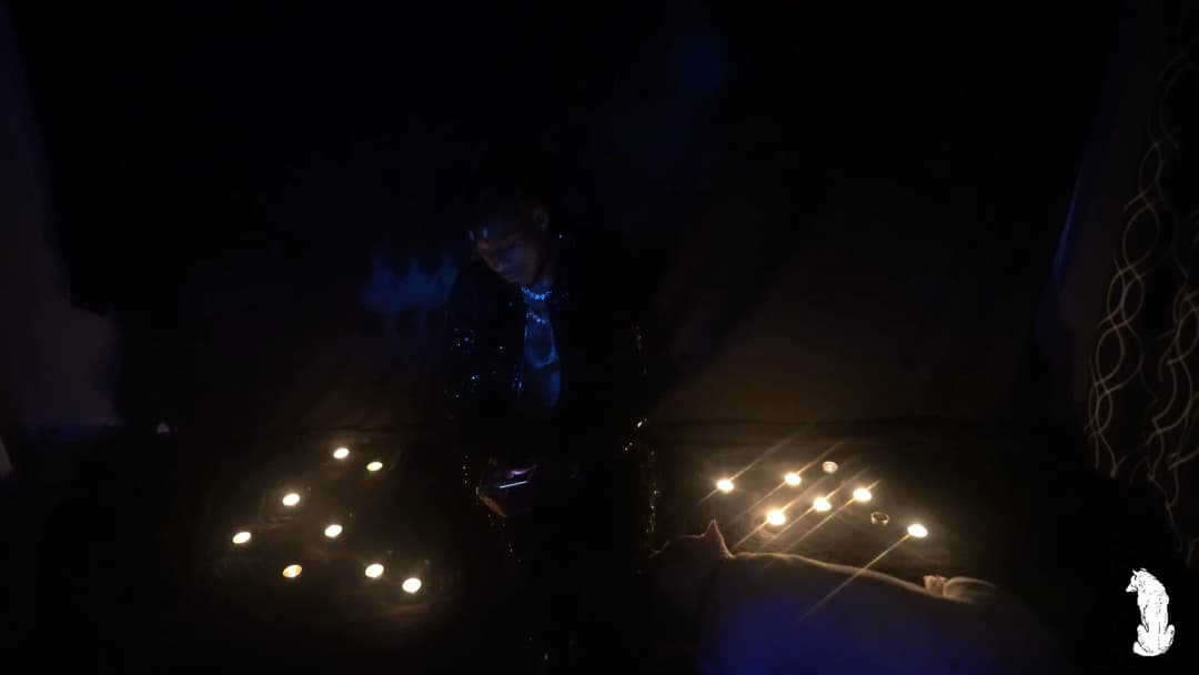 Alpha The Wolf standing between the floor candles in a dark jacket, gaze aimed downward at something just below frame, blue chain necklace glowing softly, the projected mural characters barely visible in the upper background through the haze