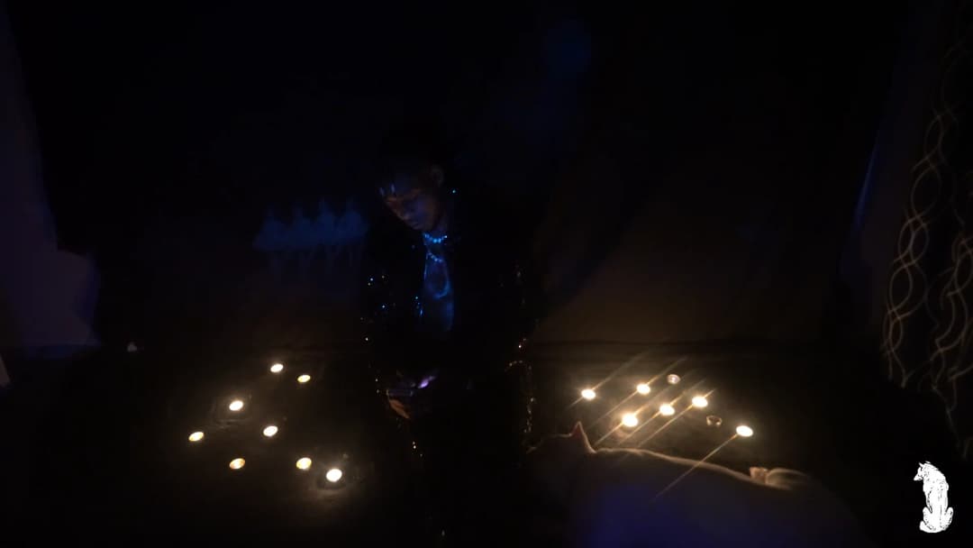 Alpha The Wolf standing in the candle circle, head tilted downward with braids catching a faint blue highlight, gaze aimed off to the left, the reflective jacket flecked with tiny points of blue light against an almost entirely black background