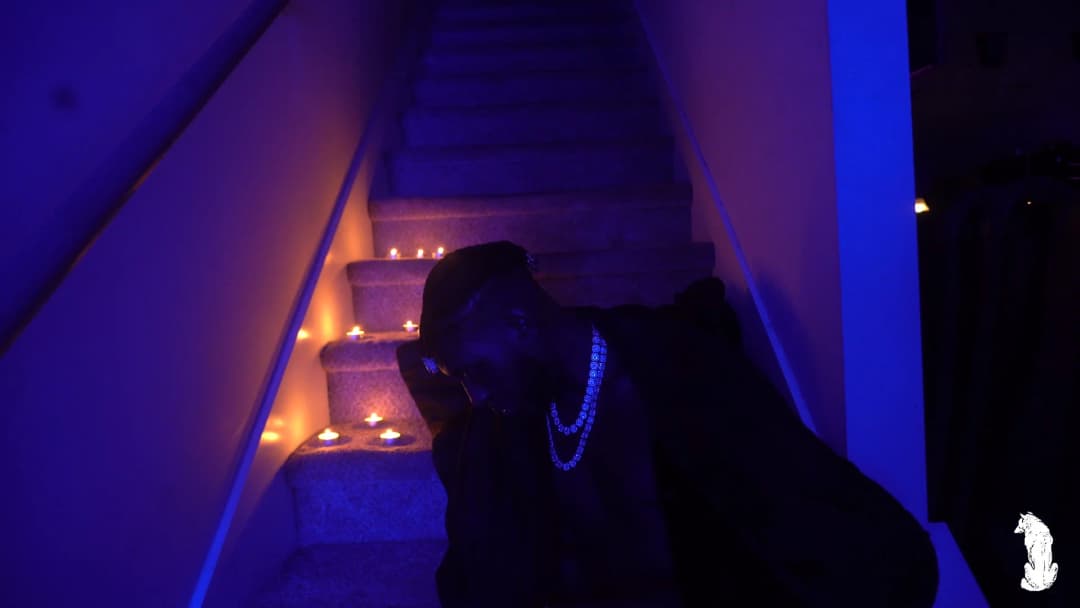 Alpha The Wolf at the base of the candle-lit staircase, face turned to look directly into the camera, hand raised to his cheek, blue iced chain hanging at the chest, expression focused and self-possessed under the blue wash of light