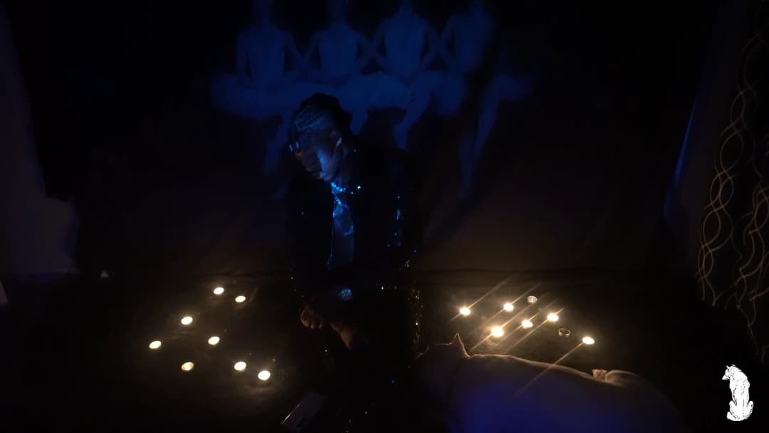 Alpha The Wolf standing over the candle clusters in a dark jacket with braids lit blue from above, looking downward with one hand loosely at his side, the faint outline of the projected mural and the symmetrical candle arrangements framing him in the wide shot