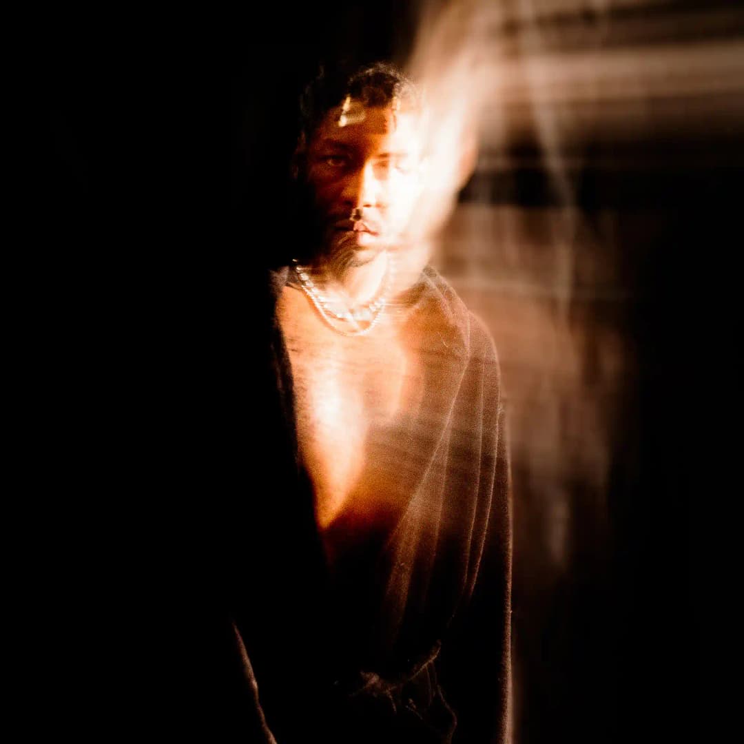 Alpha The Wolf standing in near-total darkness, lit from behind by streaks of warm white light ghosting across his chest and face, wearing a pearl necklace — a raw motion-blurred long-exposure portrait