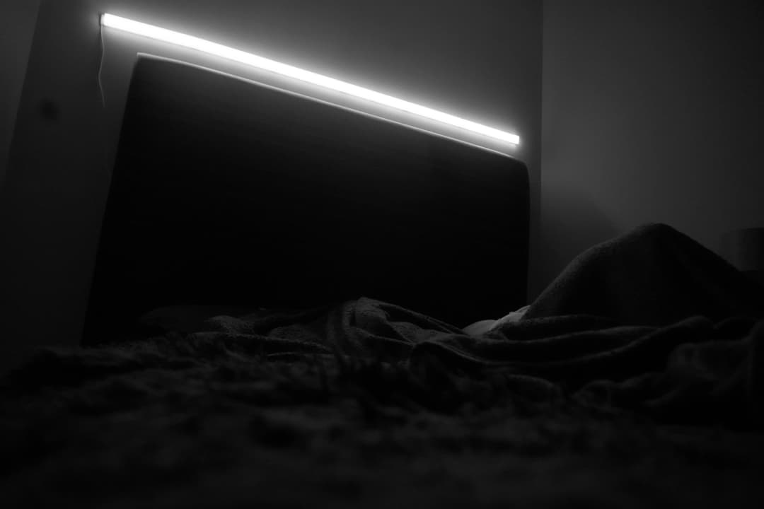Alpha The Wolf's album art depicting an empty dimly-lit bedroom in black-and-white, a single LED strip glowing above an upholstered headboard and rumpled sheets — a desolate intimate still life