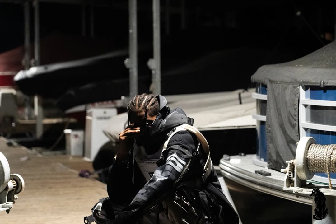 Alpha The Wolf standing at a nighttime marina dock in tactical outerwear with braided hair, boats and dock equipment in the background