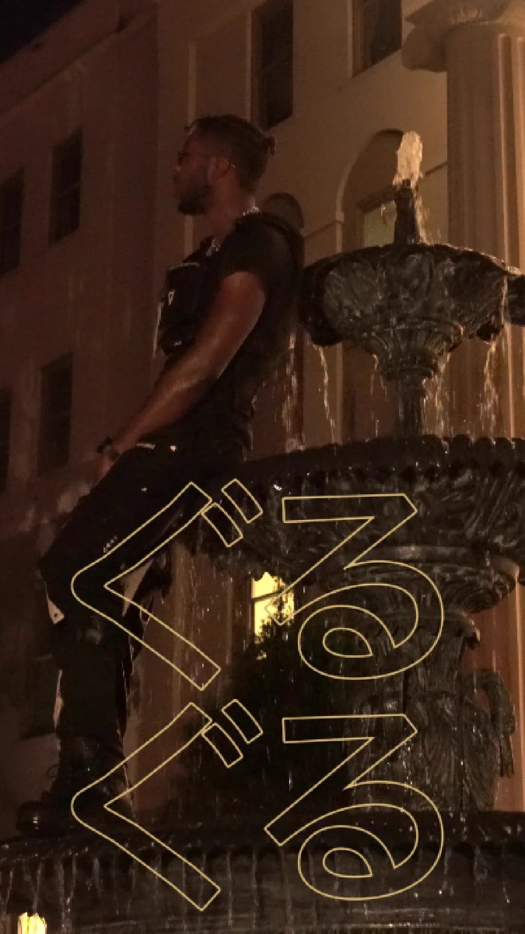 Alpha The Wolf standing shirtless on the basin of an ornate multi-tiered fountain at night, lit by warm ambient light, with stylized title text overlaid