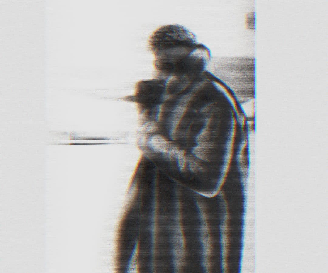 Alpha The Wolf standing and cradling a camera close to the face, wearing glasses and a coat, in a close-cropped grainy portrait with soft ghosting