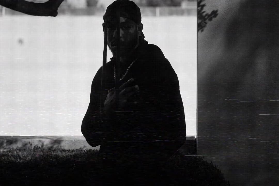 Alpha The Wolf in silhouette facing camera, arms crossed with hand on chest, chain necklace catching light — datamoshed glitch on bright outdoor backdrop