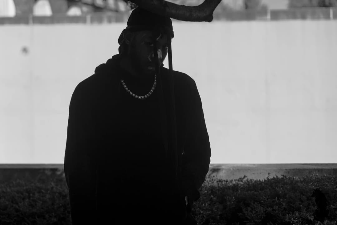 Alpha The Wolf standing outside near a bare tree branch, dark hooded jacket and chain necklace against a light concrete wall — clean raw shot