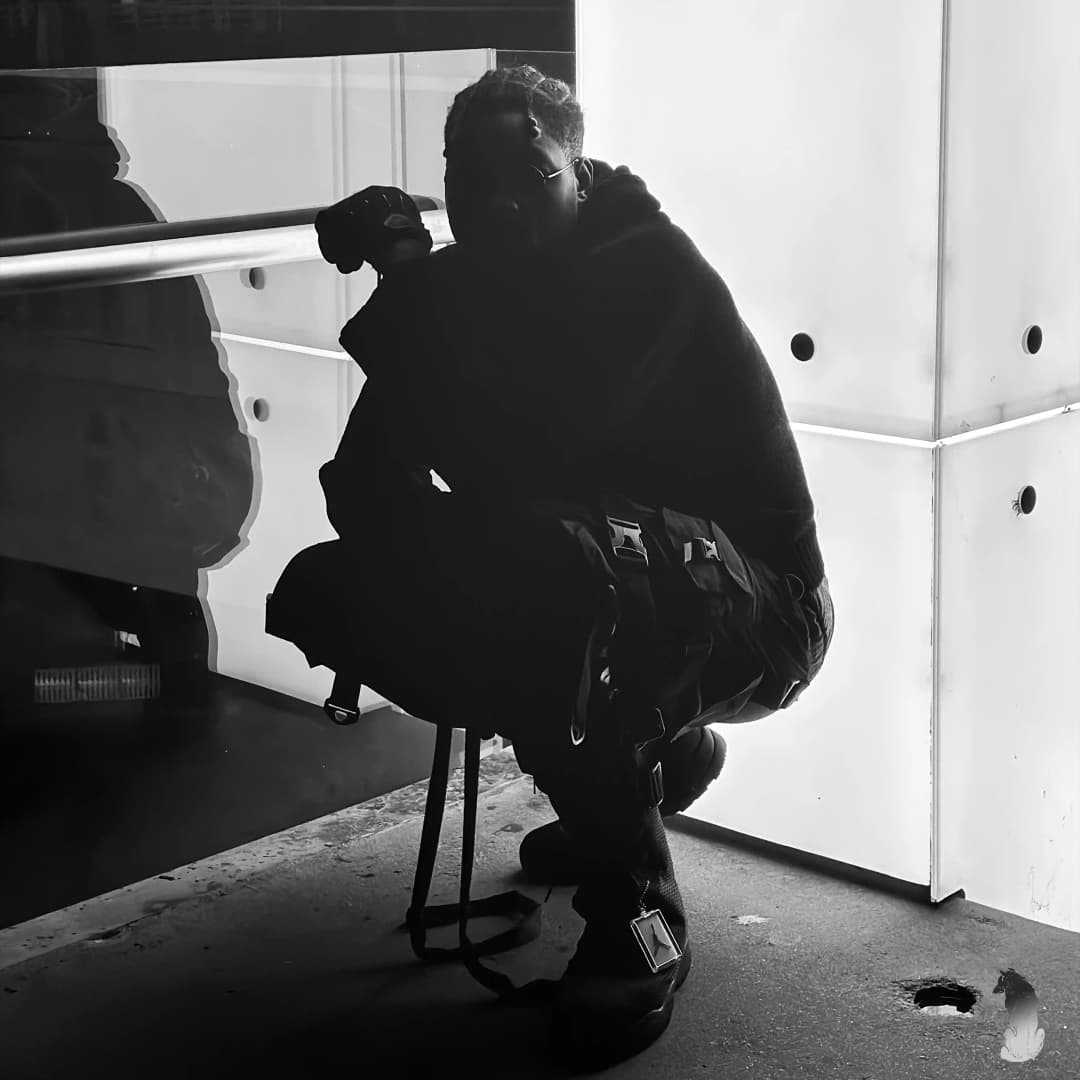 Alpha The Wolf crouching low in all-black clothing on a concrete surface with a large mirror panel and white wall behind him — raw base photo for the album artwork