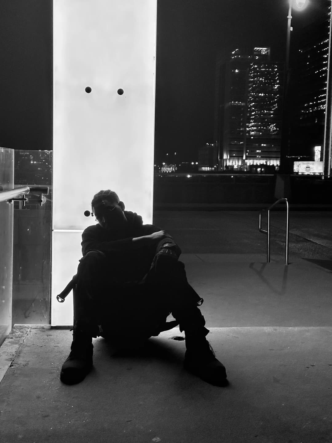 Alpha The Wolf seated on concrete ground at the base of a glowing white architectural column, knees pulled up and head resting in one hand, black-and-white frame with a lit downtown skyline visible across an open plaza behind him
