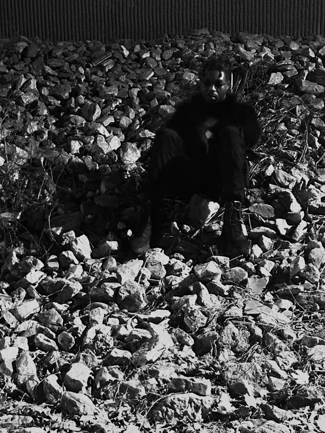 Alpha The Wolf seated among a field of broken rubble and crushed rock outdoors, dressed in all black, body folded inward with hands clasped, shot in high-contrast black and white against a corrugated metal wall