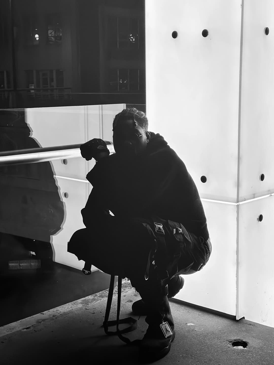 Alpha The Wolf perched on a low stool in a deep squat beside a luminous white architectural panel, face tilted upward toward the light with one hand raised near his chin, his reflection faintly visible in the dark glass behind him, black and white