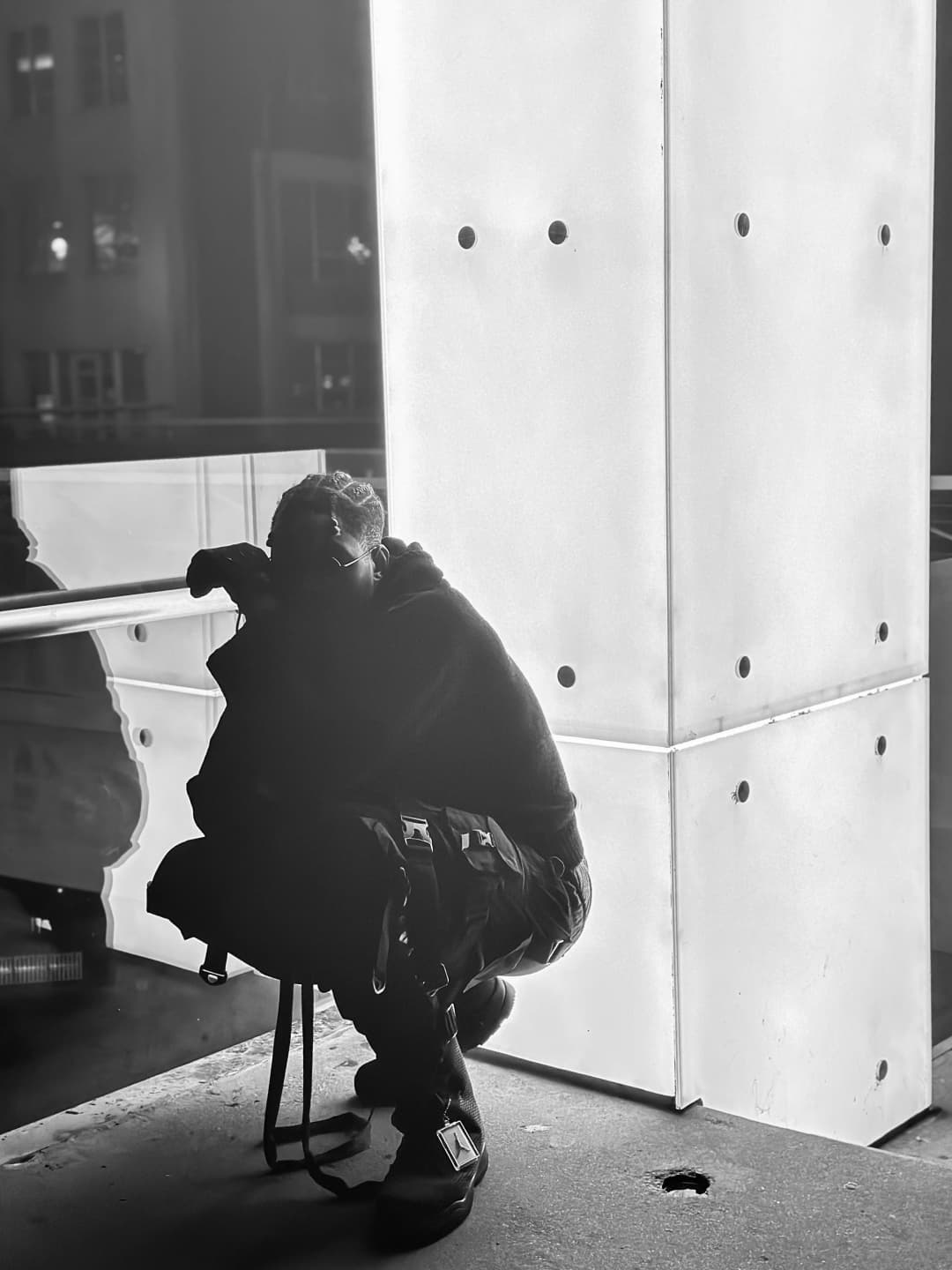 Alpha The Wolf seated on a stool with his back to the camera, hunched forward and head bowed toward the glowing white panel beside him, the downtown streetscape reflecting faintly in the dark glass to his left, shot in black and white