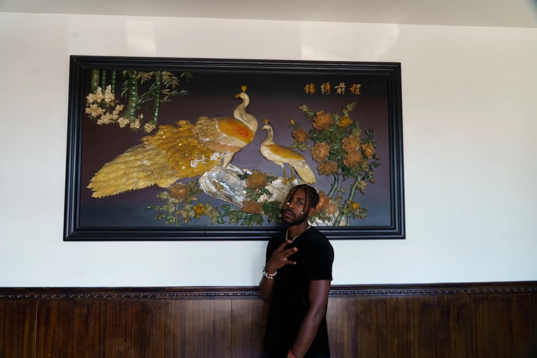 Alpha The Wolf throwing up a hand sign while standing beneath a large ornate Asian painting of golden peacocks, wearing a black tee and chain jewelry, the painting's dark lacquered frame and wood-paneled wainscoting of a Chinese restaurant dining room surrounding him