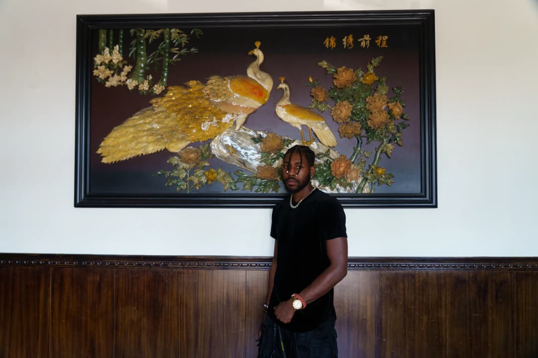 Alpha The Wolf standing slightly to the side beneath the peacock painting, body angled with a cool slouch, pearl necklace and gold watch catching the light, looking just off-camera with a relaxed confidence, the restaurant's dim natural light creating a flat, editorial tone