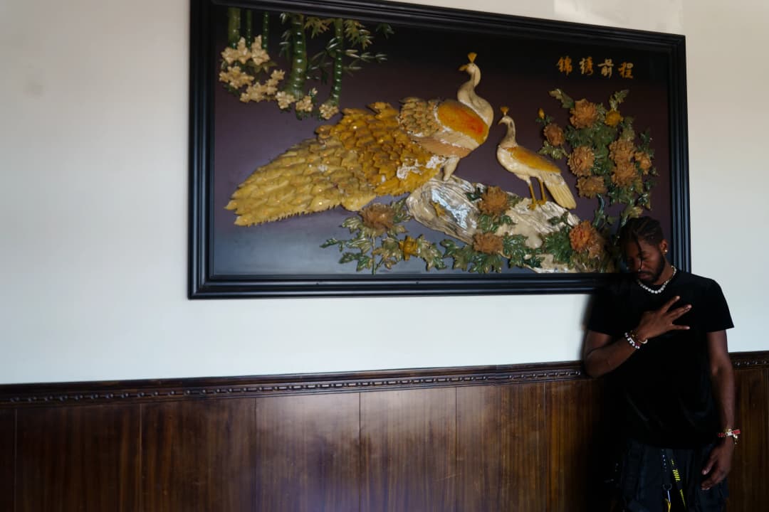 Alpha The Wolf with his hand pressed to his chest in a sincere gesture beneath the peacock painting, head slightly bowed, wearing a black tee and chain bracelet, the warm-toned restaurant interior giving the frame a nostalgic, low-key intimacy