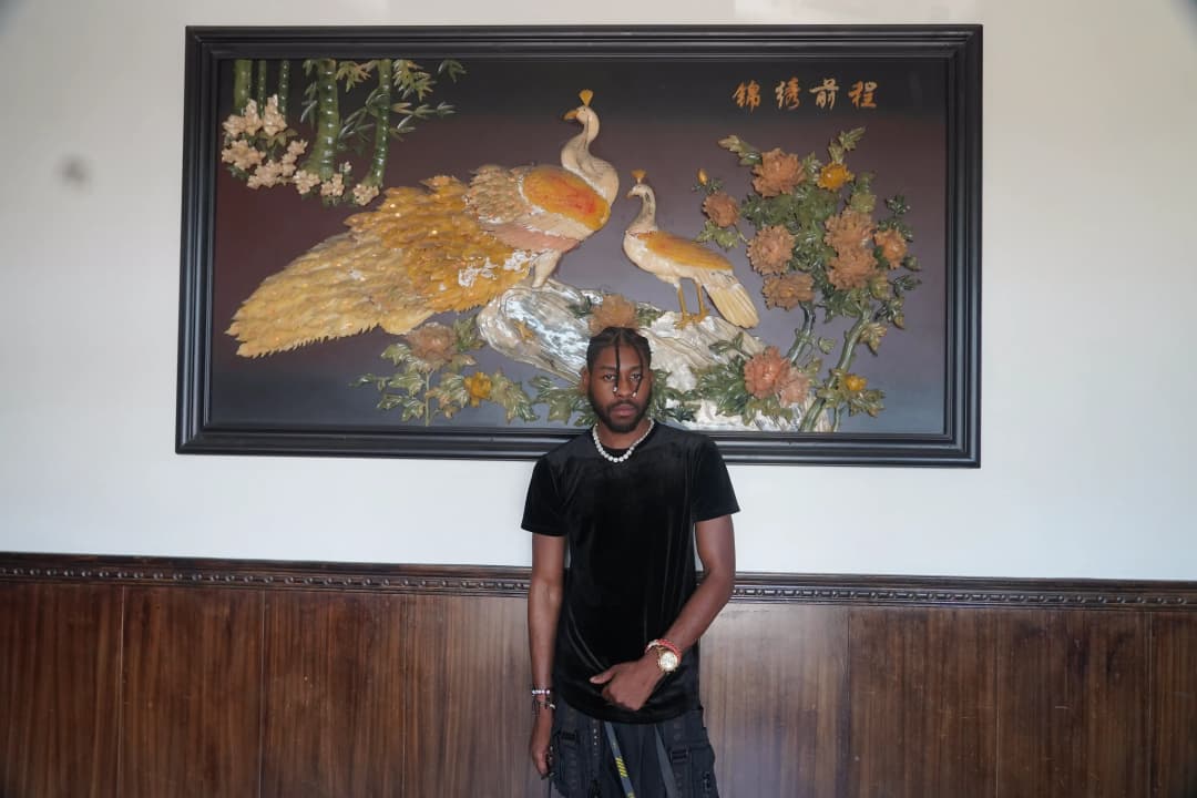 Alpha The Wolf standing full-length under the peacock artwork in a black tee and dark cargo pants, the panel Chinese characters visible at its upper corner