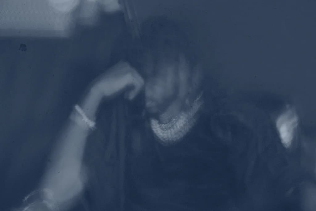 Alpha The Wolf photographed in a dimly lit interior, head bowed and hand raised near the face, shot through a translucent surface in a heavy cool blue monochromatic wash