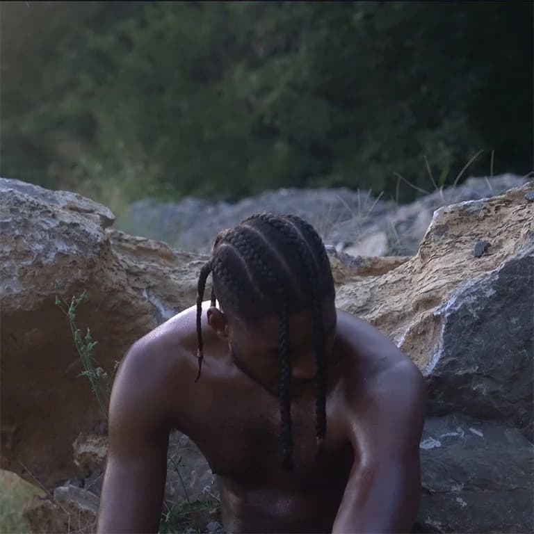 Alpha The Wolf shirtless with braids, head bowed and face angled down between large weathered boulders with green forest behind him — raw, no text