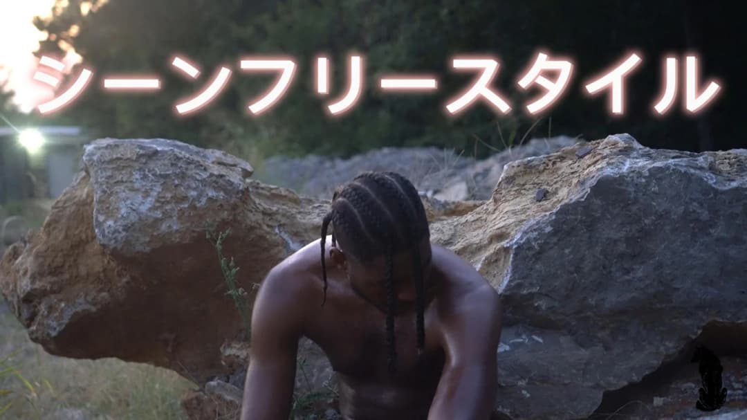 Alpha The Wolf shirtless with braids, crouching low between large rocks in a sun-dappled clearing — wider cinematic crop with Japanese title text overlaid