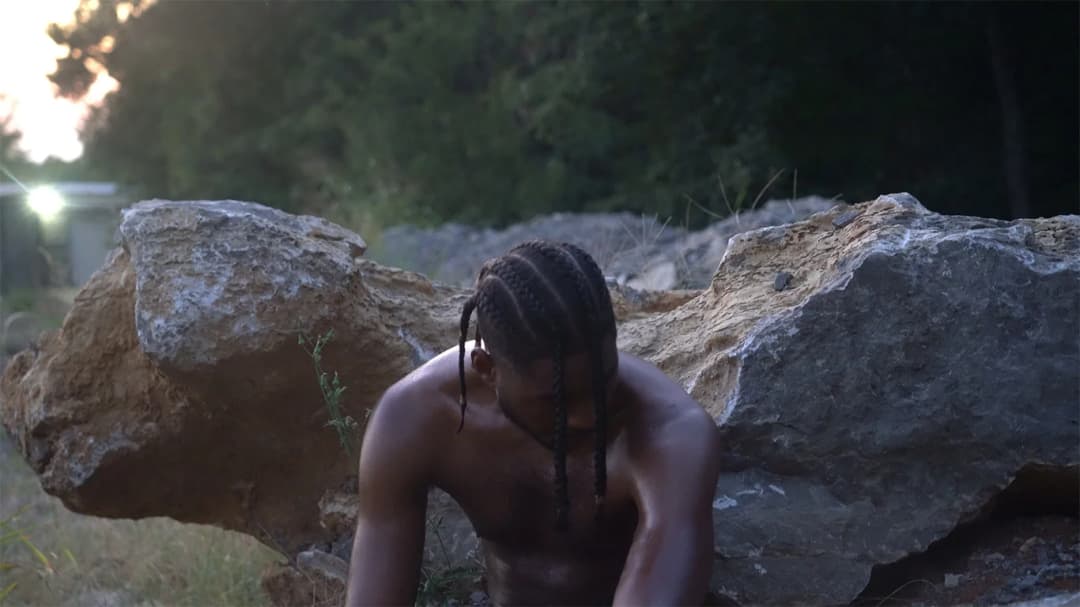 Alpha The Wolf shirtless with braids, crouching low between large rocks in a sun-dappled clearing — wider cinematic crop, no text