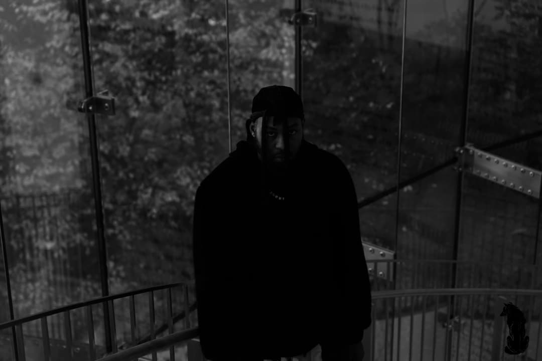 Alpha The Wolf standing inside a glass stairwell enclosure at night, trees visible through frosted panels, chain necklace, facing the camera directly in high-contrast black and white