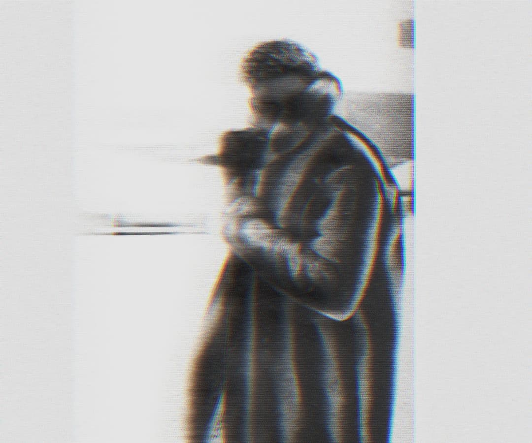 Alpha The Wolf standing and cradling a camera close to the face, wearing glasses and a coat, in a close-cropped grainy portrait with soft ghosting