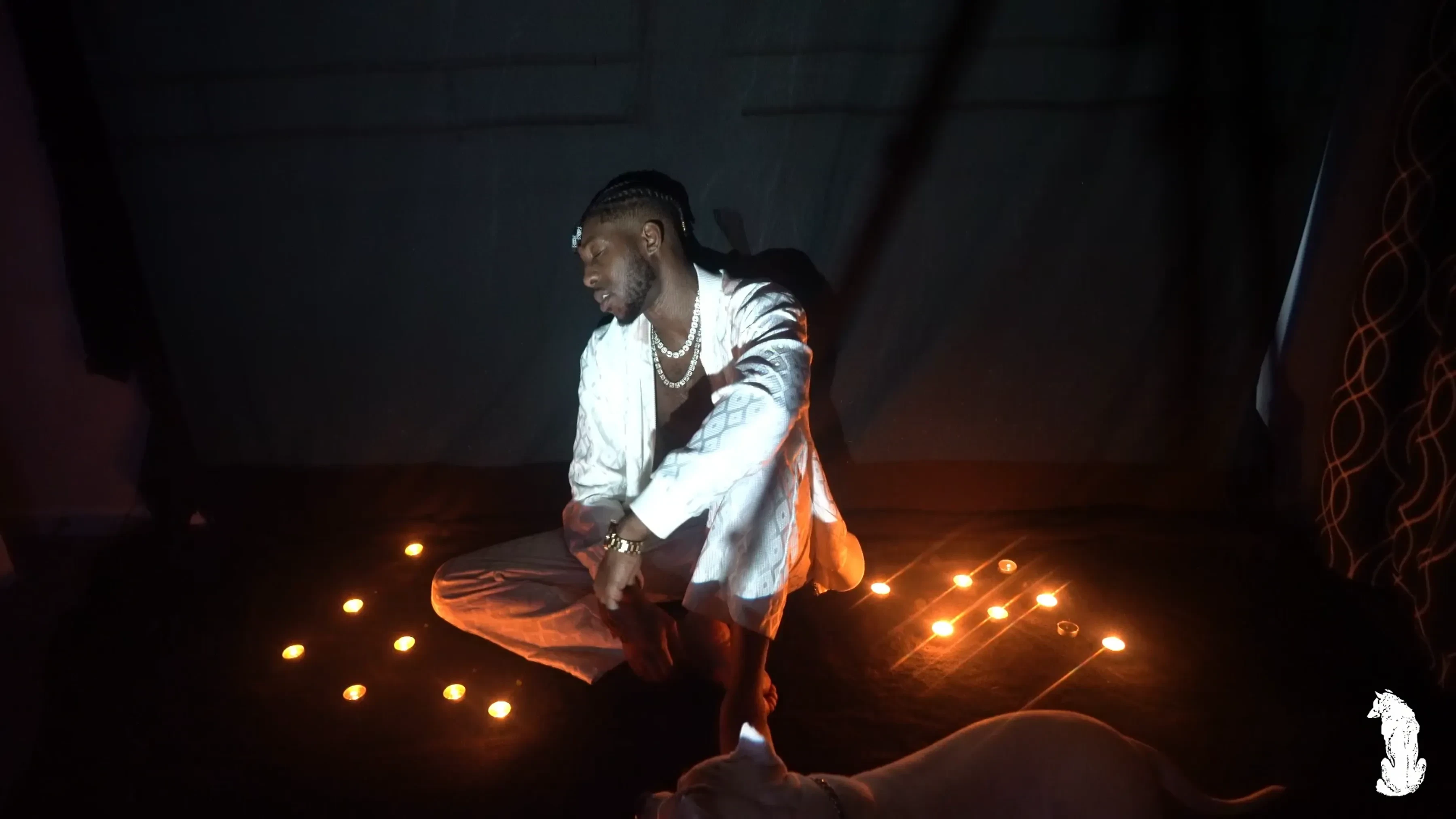 Alpha The Wolf seated cross-legged at the center of a candlelit floor in a white outfit, pearl necklace and watch catching the light, head bowed forward in stillness.