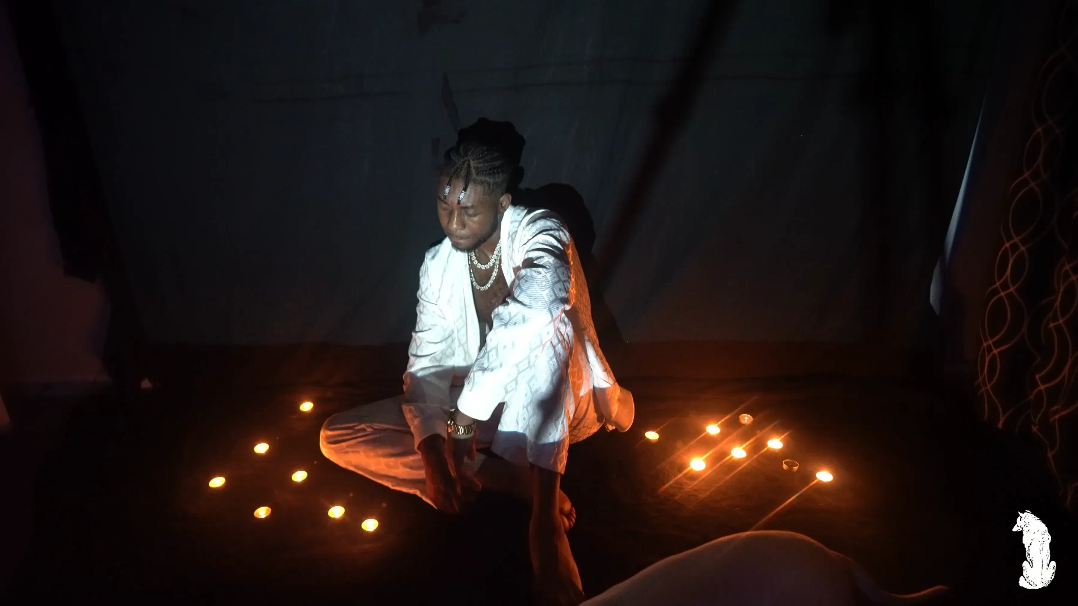 Alpha The Wolf seated at the center of the candle ring in white, face lifted toward a strong overhead spotlight, pearl necklace glowing, bright against the surrounding darkness.