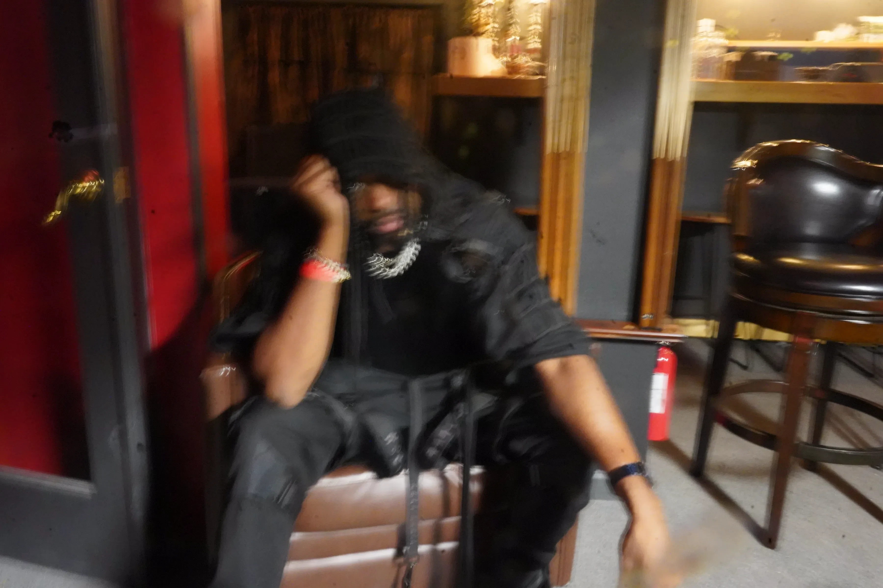 Alpha The Wolf seated low in a leather armchair in a dim bar, dressed head to toe in black with a hood up and a heavy Cuban chain, one hand raised to his face, the frame smeared by a long exposure