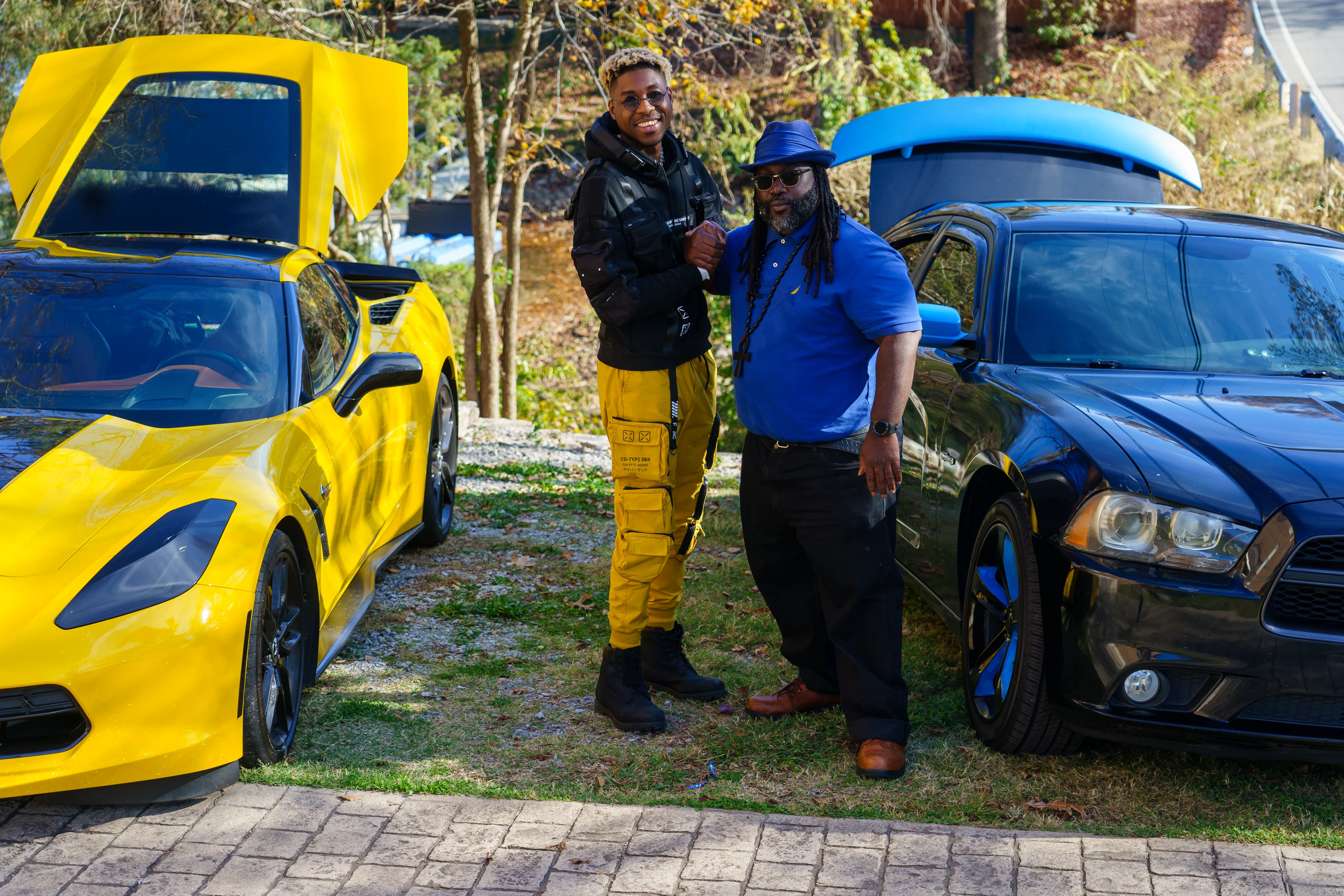 Alpha The Wolf standing with an older man in a blue polo and wide-brim hat between a yellow Corvette and a black Dodge Charger on a driveway surrounded by autumn trees, both gripping hands in a relaxed stance.