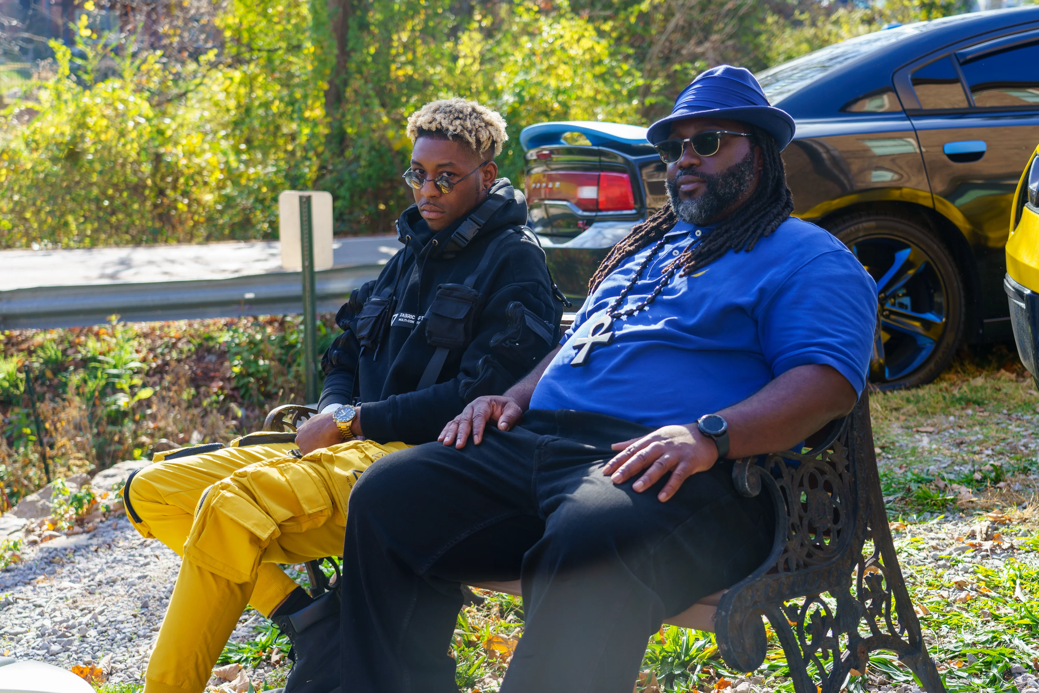 Alpha The Wolf seated on an ornate iron bench beside an older man in a blue shirt and hat, both in sunglasses and leaning back in an unbothered pose, a yellow Corvette visible behind them.