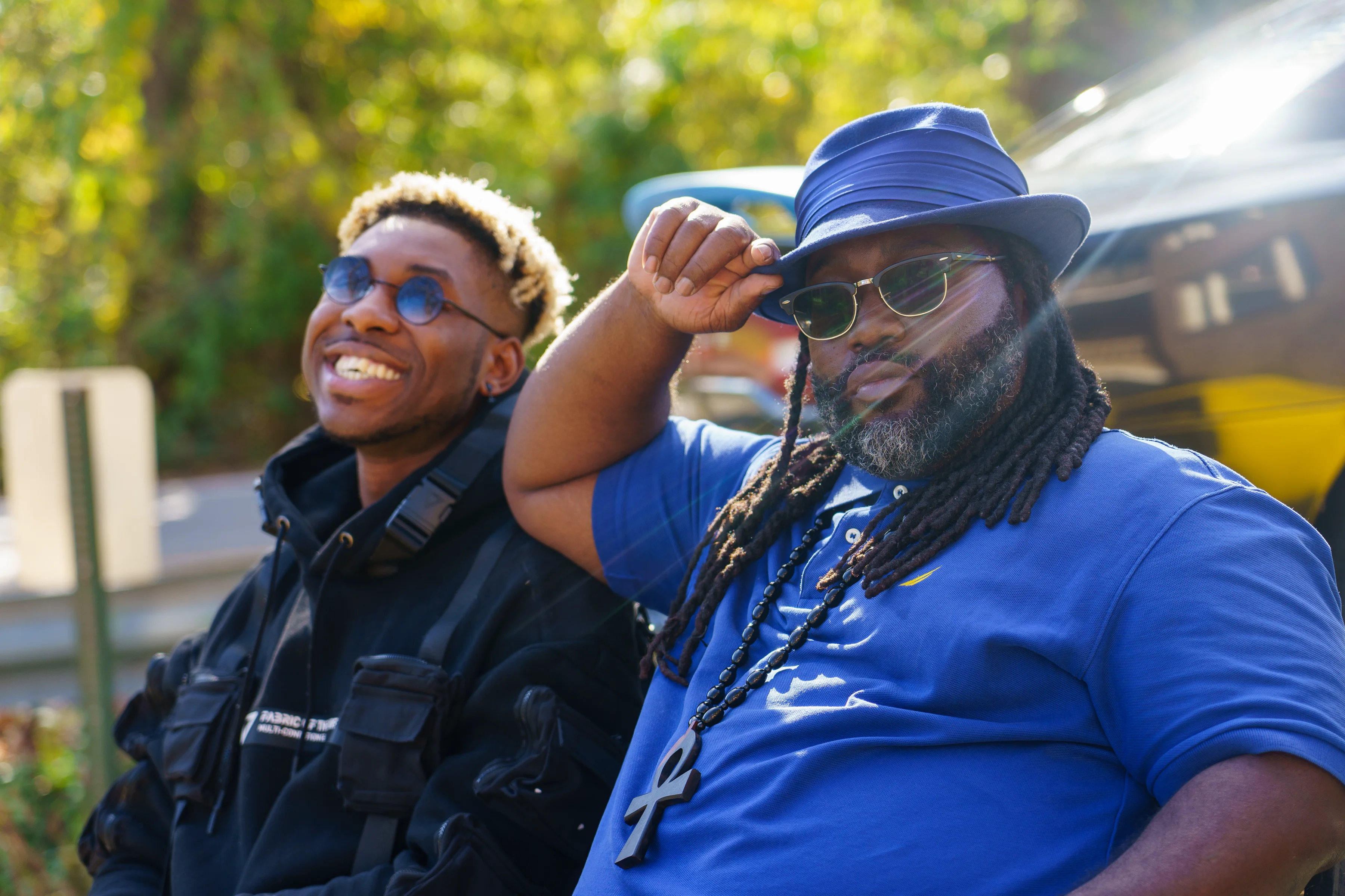 Alpha The Wolf laughing broadly beside an older man who tips his blue hat toward the camera, both in sunglasses with warm sunlight on their faces and a yellow Corvette glowing behind them.
