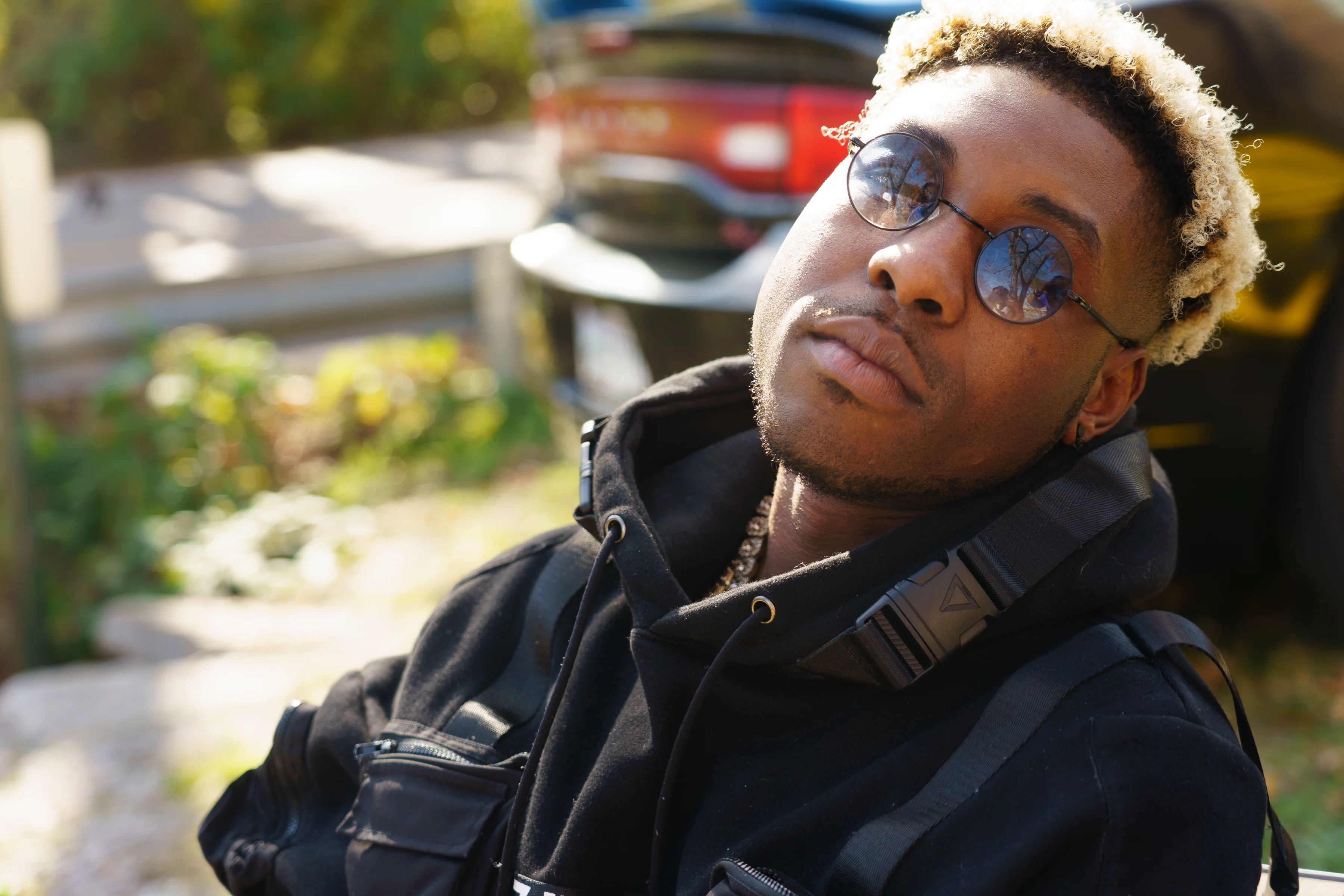 Alpha The Wolf in a close-up portrait, head tilted back slightly with a composed expression, round reflective sunglasses and a black tactical jacket, parked cars and green brush blurred behind him.