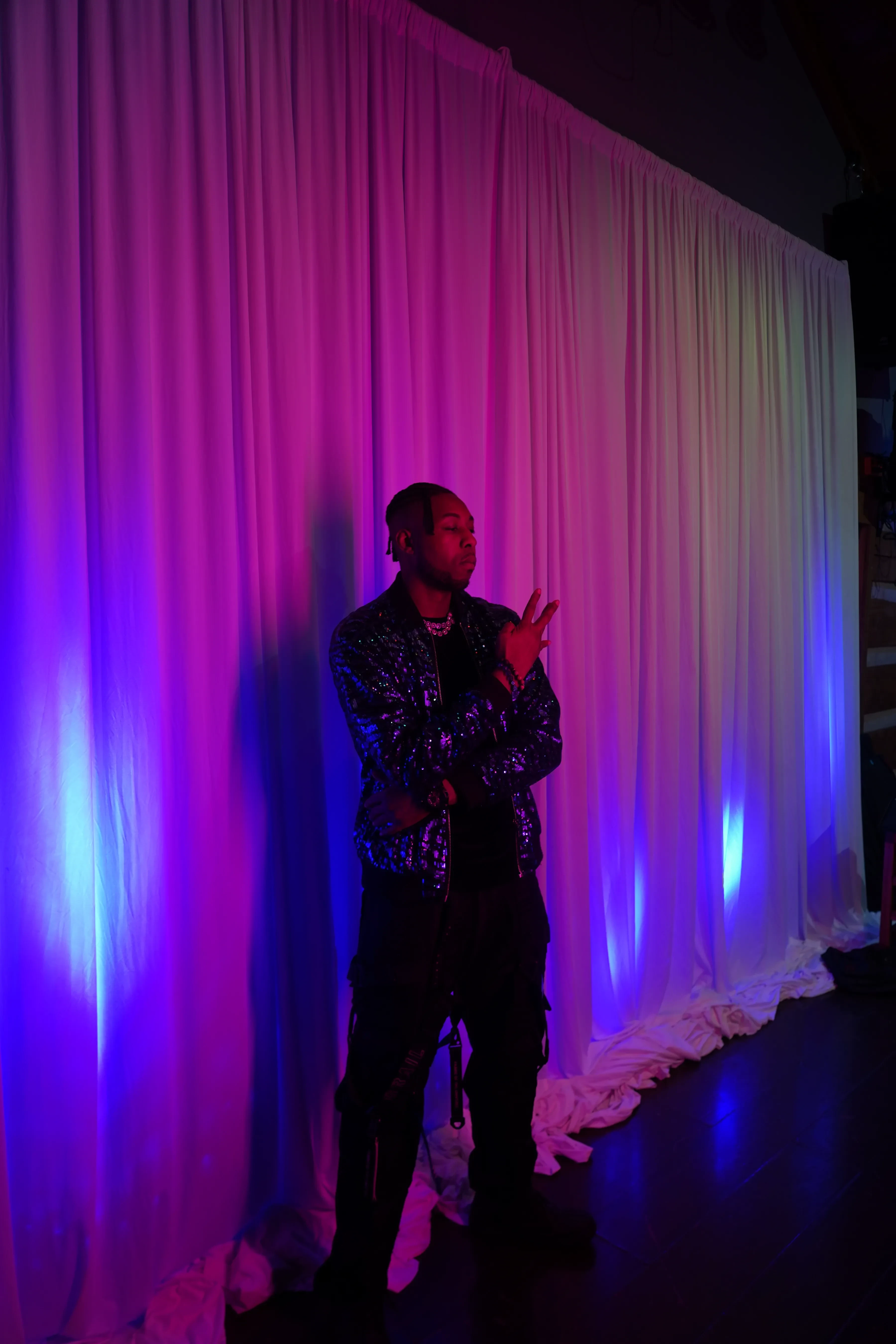 Alpha The Wolf against a white curtain flooded with magenta and blue uplight, both hands raised near his face in a split-finger gesture, the sequinned bomber catching the colour.